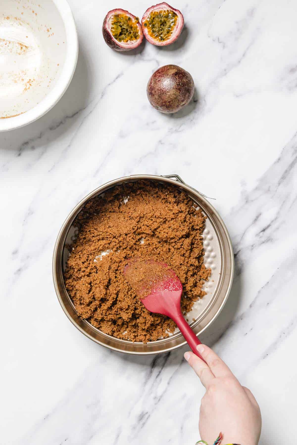 A hand spreads brown crumb mixture in a round metal pan with a red spatula, prepping the base for Passionfruit Cheesecake. Beside the pan, a halved passionfruit and a whole passionfruit rest on a white marble surface.