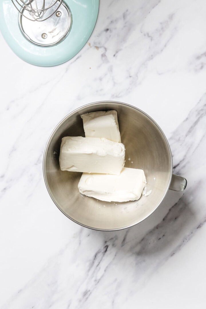 A metal mixing bowl containing three blocks of cream cheese sits on a marble surface next to part of an electric stand mixer, ready for whipping up a luscious passionfruit cheesecake.