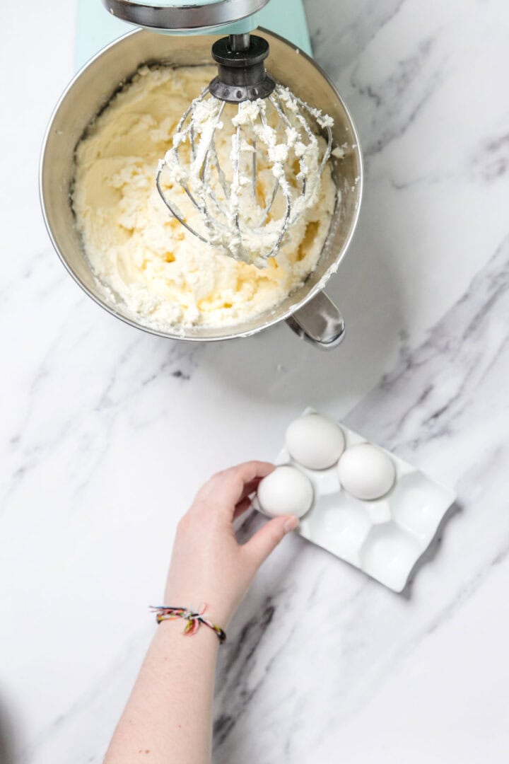 A hand reaches for an egg from a ceramic egg holder next to a stand mixer bowl filled with creamed butter and sugar—perfect for making Passionfruit Cheesecake. The whisk attachment is coated with the mixture. Marble countertop background.