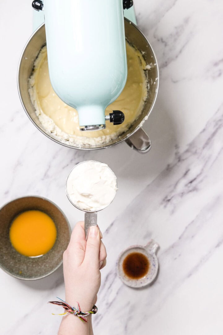 A hand holding a measuring cup of cream cheese for Passionfruit Cheesecake above a stand mixer bowl with batter. Nearby sit a bowl of melted butter and a cup of vanilla extract on a white marble surface.