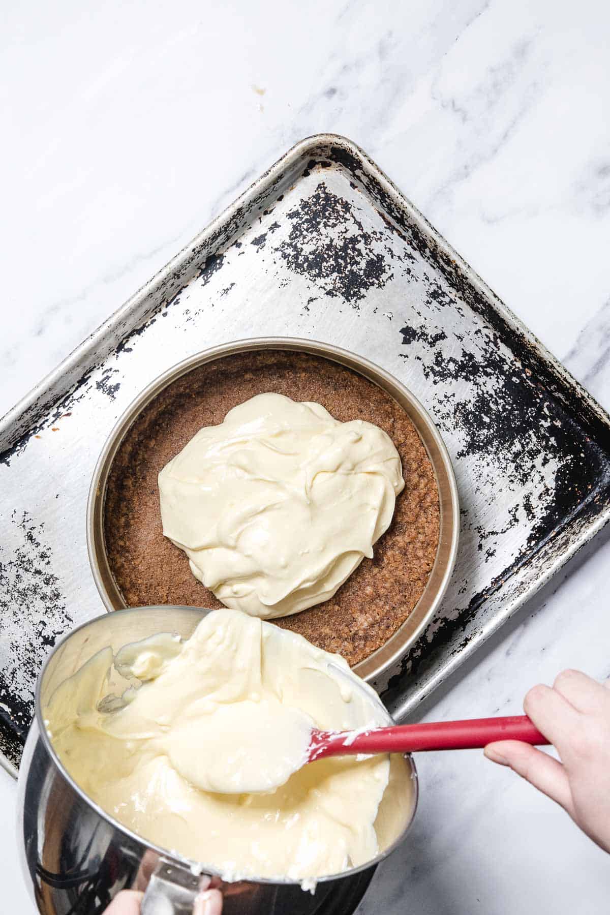 A hand uses a red spatula to spread creamy passionfruit cheesecake batter from a mixing bowl onto a crumb crust in a round pan set on a worn baking sheet.