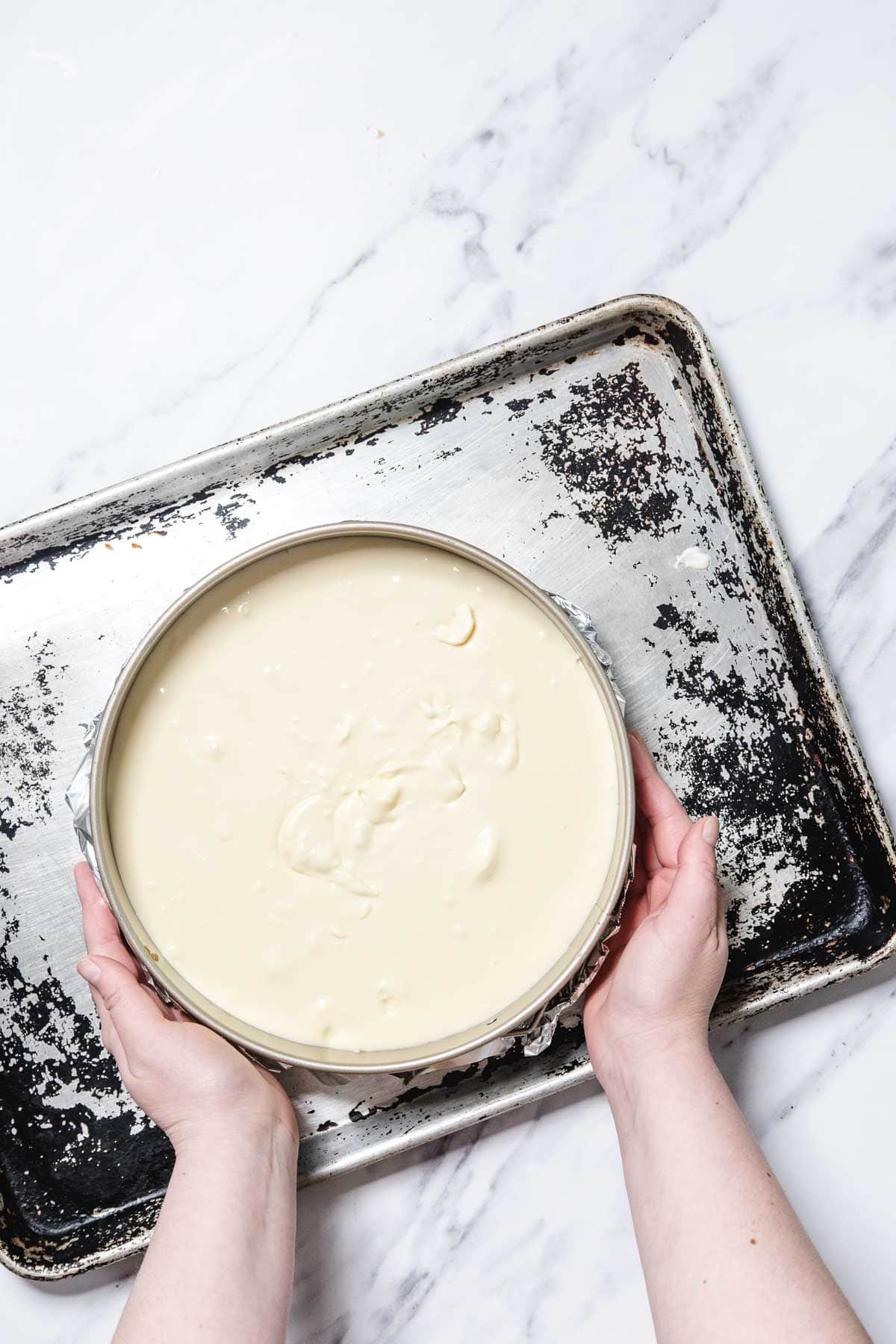 A person holding a round pan filled with uncooked passionfruit cheesecake batter, placed on a worn metal baking sheet on a white marble surface.