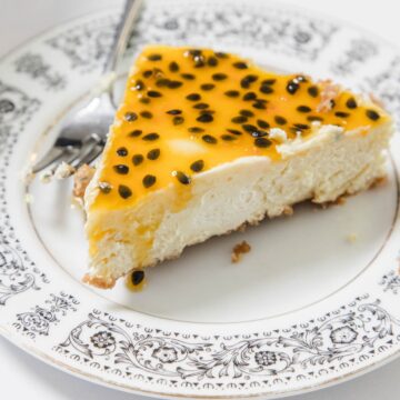 A slice of Passionfruit Cheesecake topped with tangy pulp and seeds sits on a decorative white plate with a floral pattern. A fork rests behind the creamy slice, ready to enjoy.