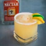 A short glass filled with a pale yellow Aguardiente Sour cocktail, garnished with a lemon and lime twist, sits on a gray surface. A bottle labeled "Aguardiente Nectar" is partially visible in the background.