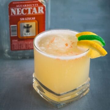 A short glass filled with a pale yellow Aguardiente Sour cocktail, garnished with a lemon and lime twist, sits on a gray surface. A bottle labeled "Aguardiente Nectar" is partially visible in the background.