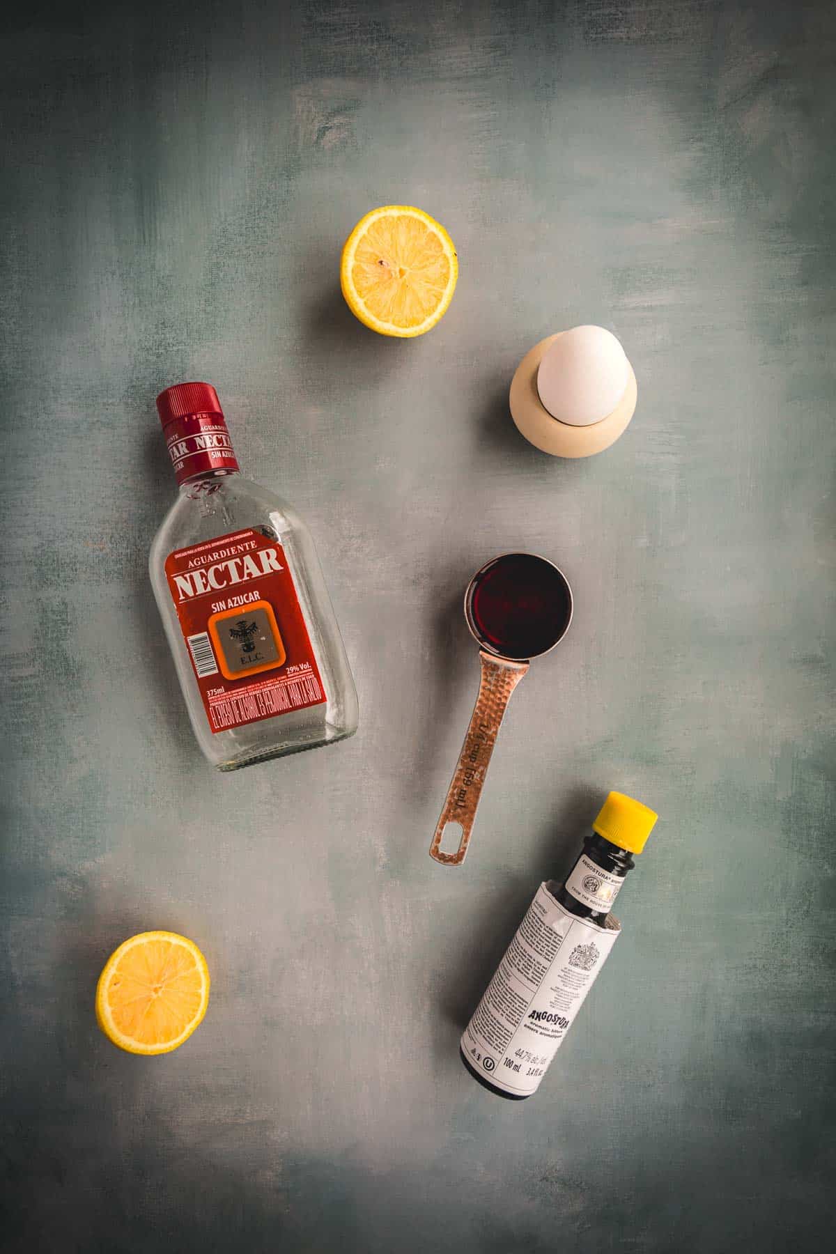 A bottle of agave nectar, a measuring cup with liquid, lemon halves, an egg in a cup, and bitters are arranged on a gray background—perfect ingredients for crafting an Aguardiente Sour, a classic Colombian cocktail.