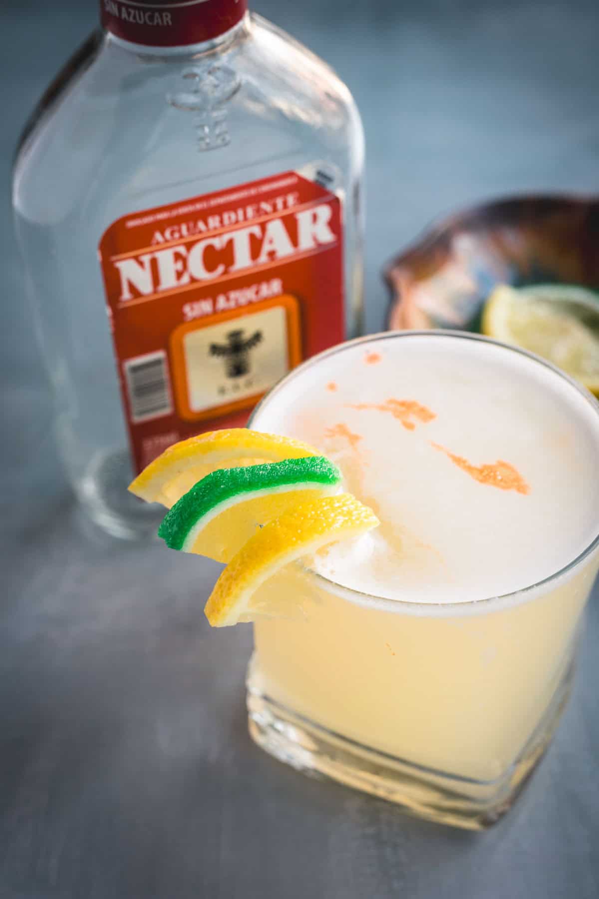 A glass of yellow Aguardiente Sour cocktail garnished with lemon and lime slices sits next to a bottle of aguardiente; a bowl with lemon wedges is in the background.