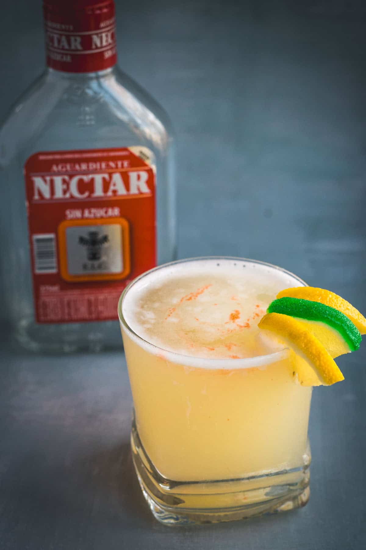 A yellow Aguardiente Sour cocktail with a frothy top is served in a short glass, garnished with lime and lemon wedges. A bottle labeled “Aguardiente Nectar” sits in the background.