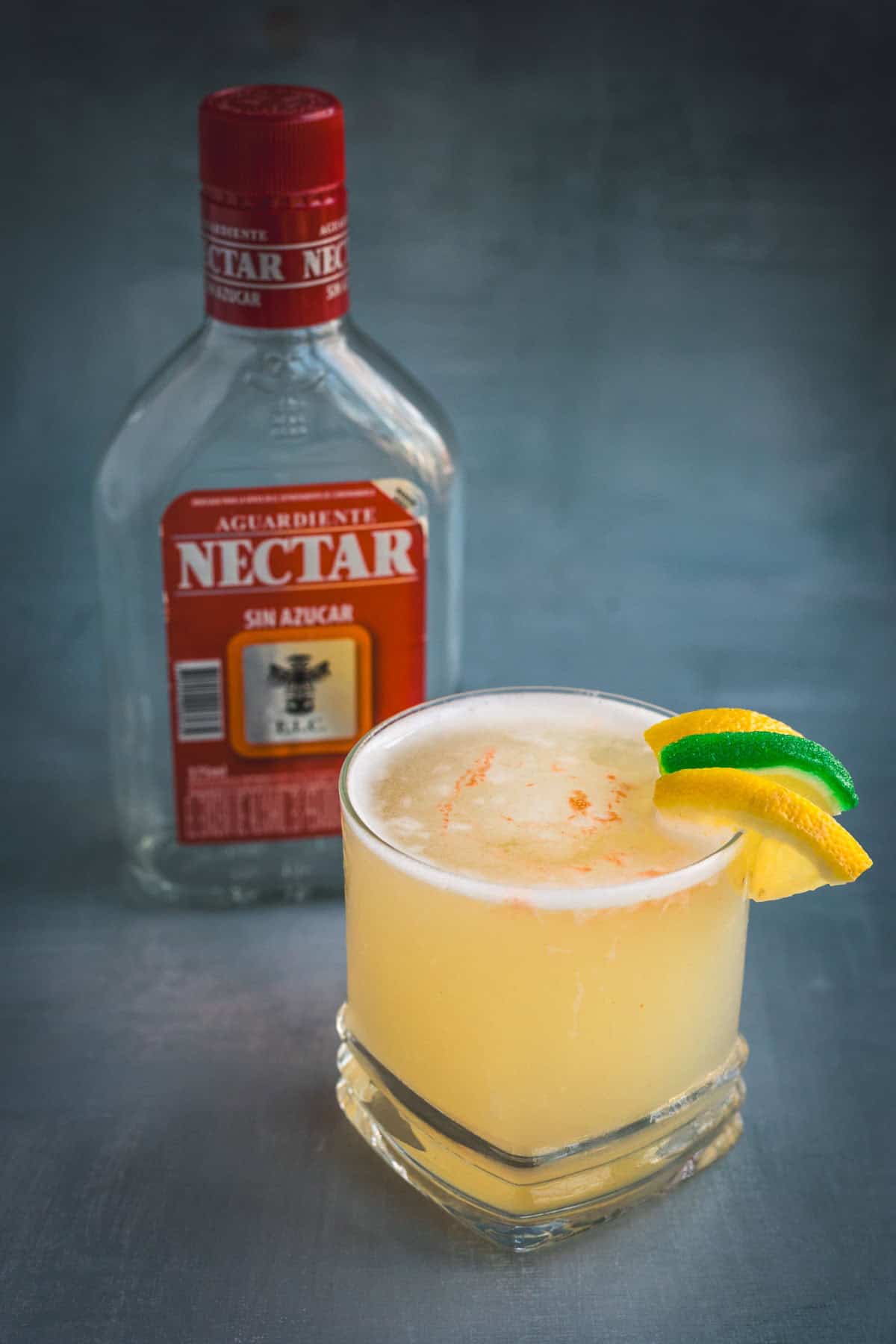 A vibrant Aguardiente Sour cocktail, garnished with lime and lemon wedges, sits before a bottle of Aguardiente Nectar with a red label on a gray surface.