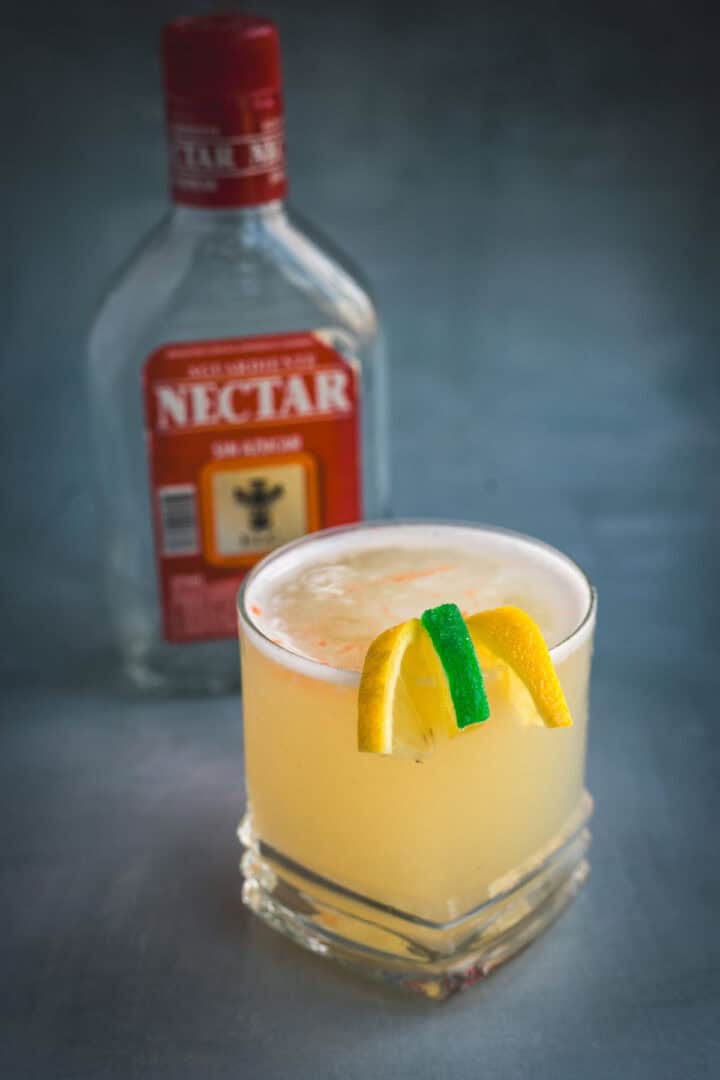 A glass of yellow Aguardiente Sour cocktail, garnished with a lemon peel, a lime peel, and an orange peel, sits in front of a clear bottle labeled “Nectar” with a red cap and label.