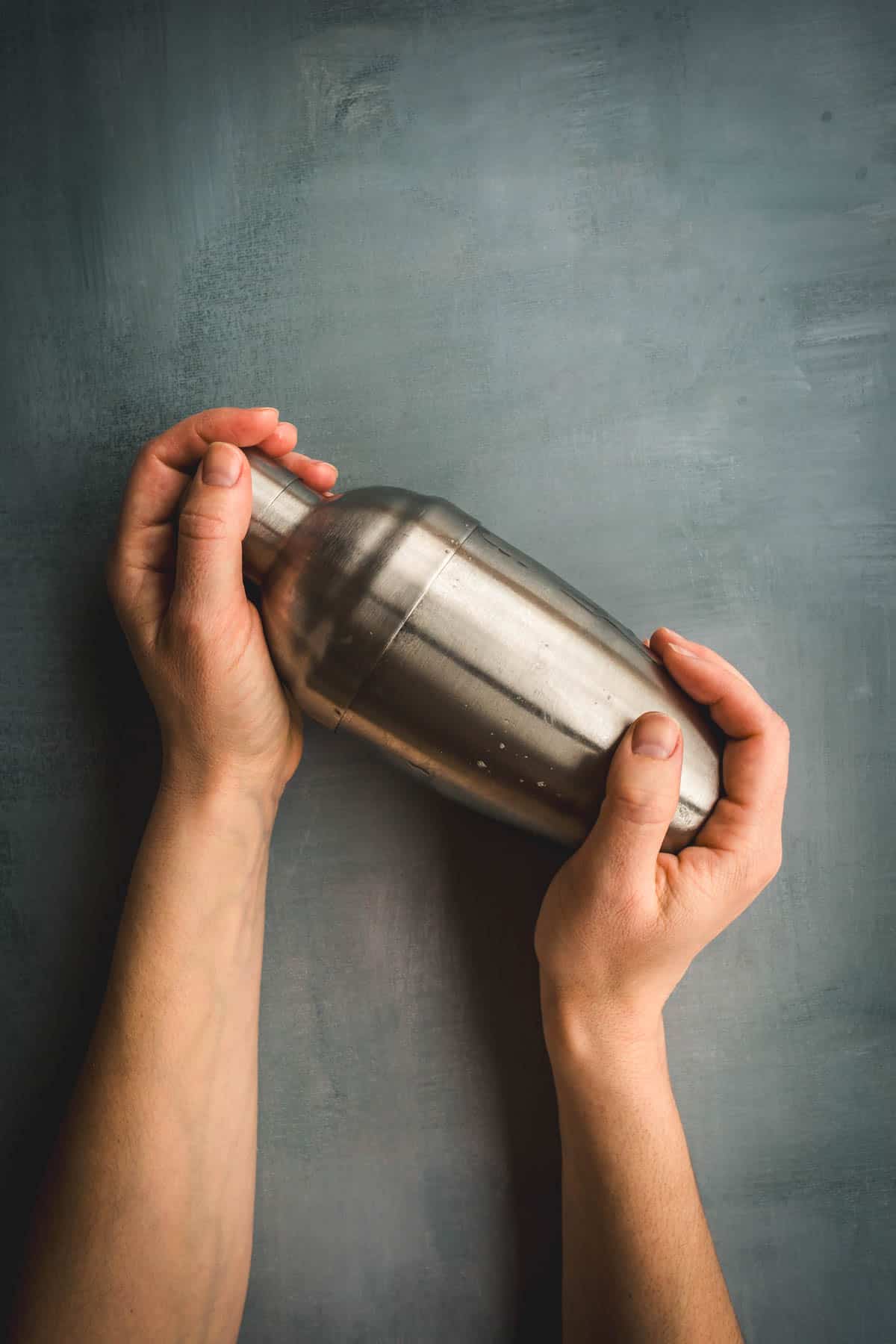A pair of hands holding a closed stainless steel cocktail shaker against a gray textured background, ready to mix an Aguardiente Sour cocktail.