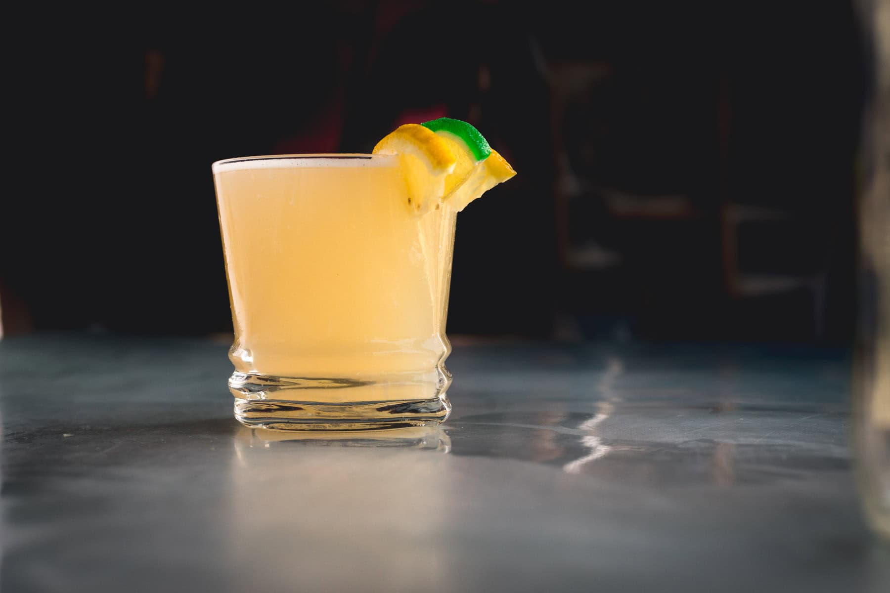 A short glass filled with a light yellow Aguardiente Sour cocktail, garnished with a lemon wedge and a lime wedge on the rim, sits on a reflective surface against a dark background.