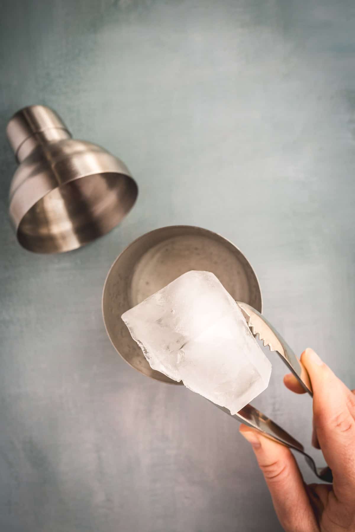 A hand holds metal tongs gripping a large ice cube above a metal cup, ready to craft an Aguardiente Sour cocktail, with a shaker cap nearby on the gray-blue surface.