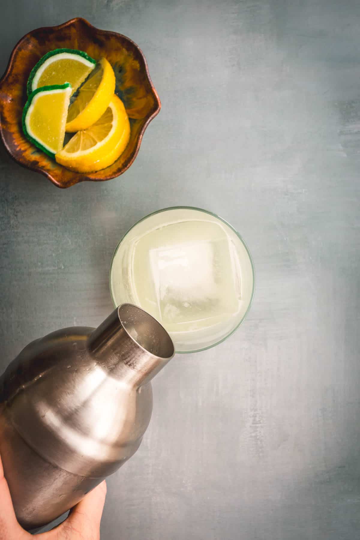 A hand holds a metal cocktail shaker, pouring an Aguardiente Sour over a large ice cube in a glass. A ceramic bowl with lemon and lime wedges sits nearby on a gray surface.