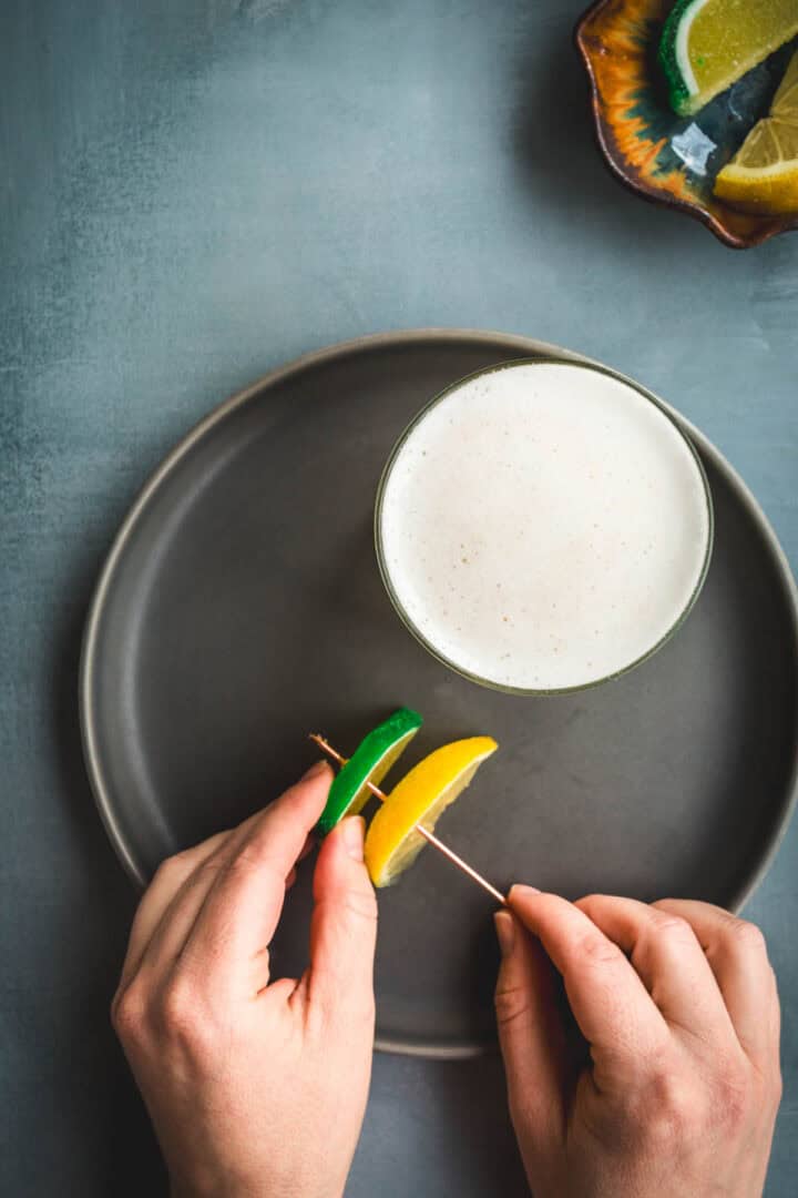 A person holds a toothpick with a lime and lemon wedge over a gray plate next to a glass of frothy Aguardiente Sour cocktail. A small bowl with more citrus wedges sits in the corner on a gray surface.