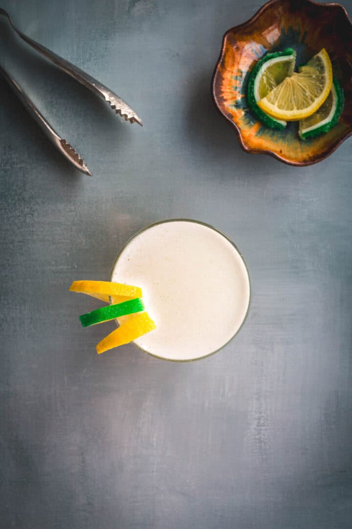 An Aguardiente Sour cocktail, frothy and garnished with lemon and lime peels, sits on a gray surface. Metal tongs rest in the top left, while a small ceramic dish with extra lemon and lime wedges is in the top right corner.