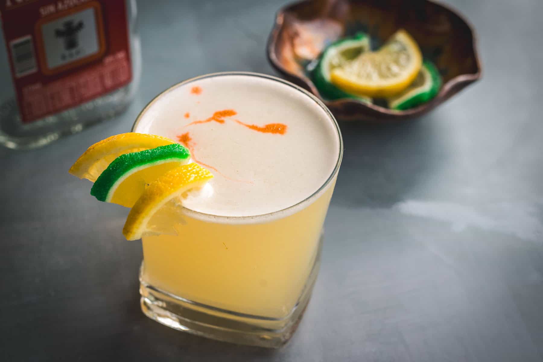 A glass of yellow Aguardiente Sour cocktail garnished with lemon and lime slices sits on a gray surface, with a bottle in the background and a small bowl of citrus slices nearby.