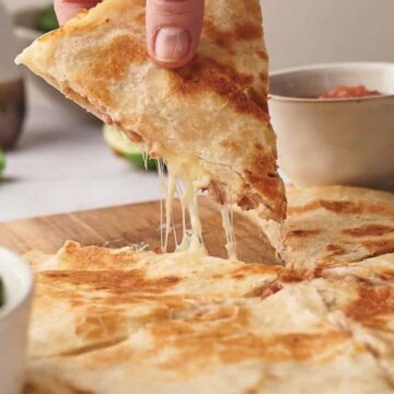 A hand lifts a triangular slice of cheesy quesadilla from a wooden board, with melted cheese stretching. Sliced quesadilla pieces and bowls of cilantro and salsa are also visible in the background.