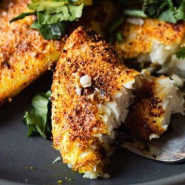 A close-up of seasoned, baked white fish fillets on a dark plate with a fork breaking off a piece. Fresh cilantro leaves are sprinkled on top and around the fish.
