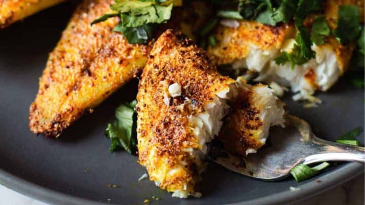 A close-up of seasoned, baked white fish fillets on a dark plate with a fork breaking off a piece. Fresh cilantro leaves are sprinkled on top and around the fish.