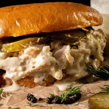 A close-up of a chicken sandwich with creamy white sauce, sliced pickles, and a brioche bun, resting on parchment paper with sprigs of dill, peppercorns, and a jar of sauce in the background.