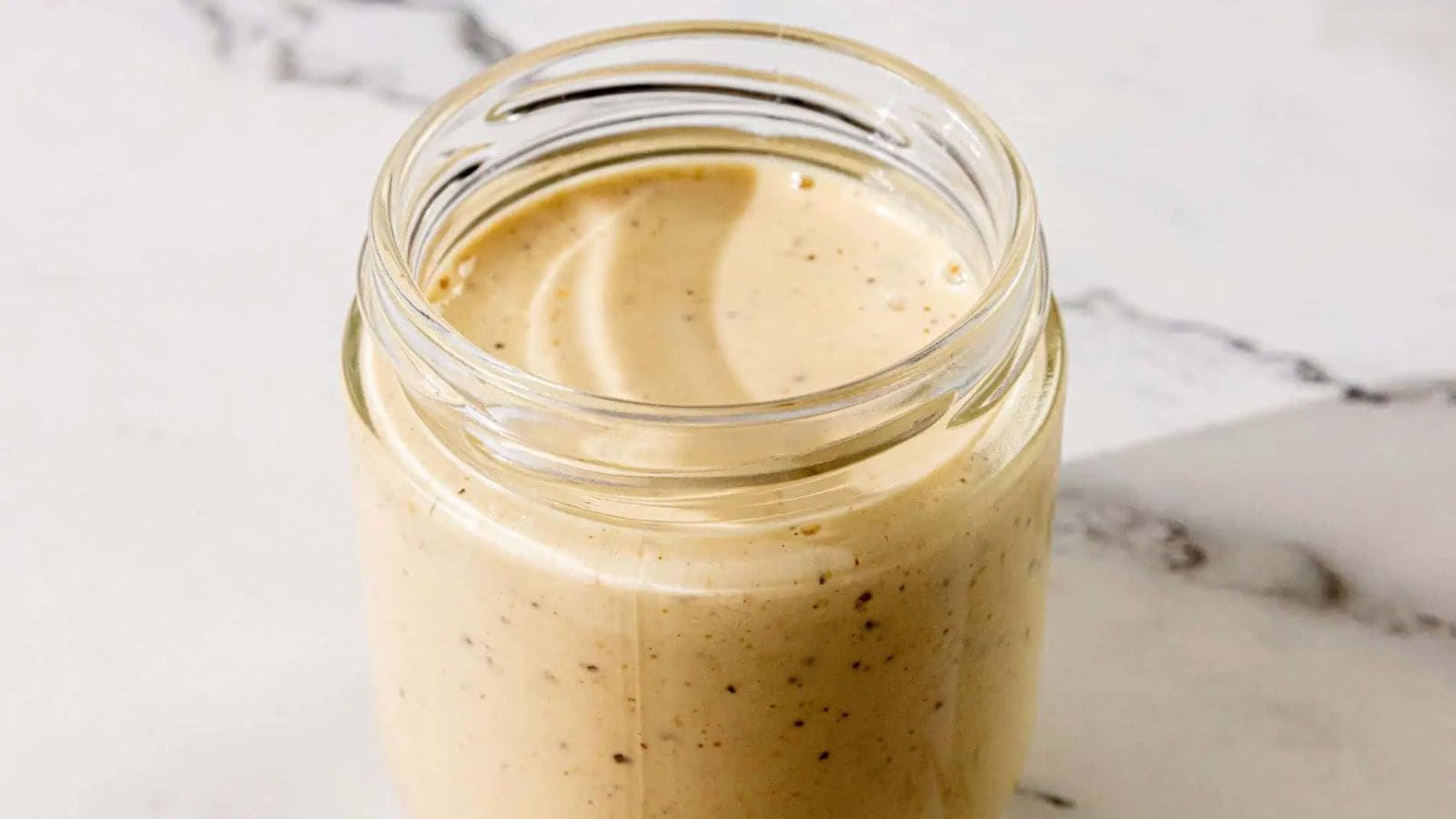 A glass jar filled with creamy, light brown Alabama White Sauce, speckled with black pepper, sits on a white marble surface—perfect for drizzling over chicken or as a tangy BBQ sauce.