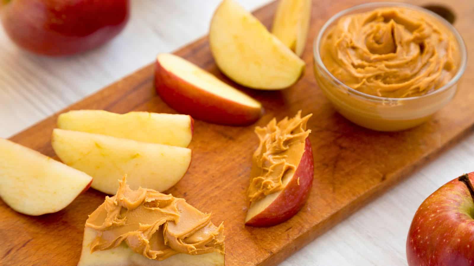 Apple slices arranged on a wooden board, some topped with peanut butter. A small glass bowl filled with peanut butter is next to the apples. Whole apples are also visible in the background.
