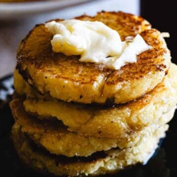 A stack of four golden brown cornmeal pancakes sits on a dark plate, topped with a dollop of melting butter.