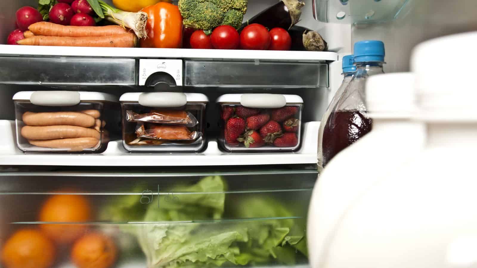 A refrigerator filled with fresh vegetables, sausages, packaged food, strawberries in containers, a bottle with a blue cap, and partially visible milk jugs in the foreground.