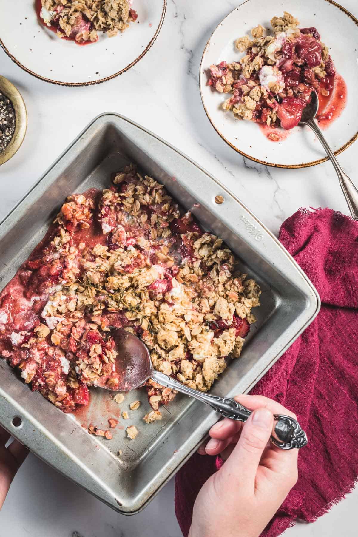Balsamic Strawberry Crisp With Goat Cheese : At the Immigrant's Table 30 A hand holds a metal baking dish containing a partially served Balsamic Strawberry Crisp with a spoon. Two plates with portions of the crisp and spoons are visible on a white marble surface, along with a red cloth napkin.