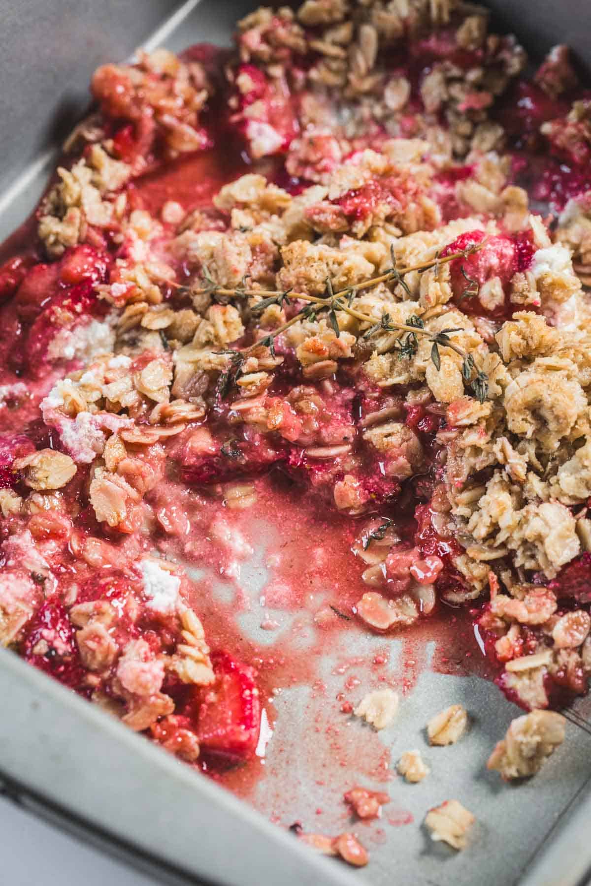 Balsamic Strawberry Crisp With Goat Cheese : At the Immigrant's Table 16 A close-up of a partially eaten Balsamic Strawberry Crisp in a baking dish, showing oats and crumb topping over a juicy, red fruit filling with a sprig of thyme on top—a perfect strawberry dessert for balsamic recipes lovers.