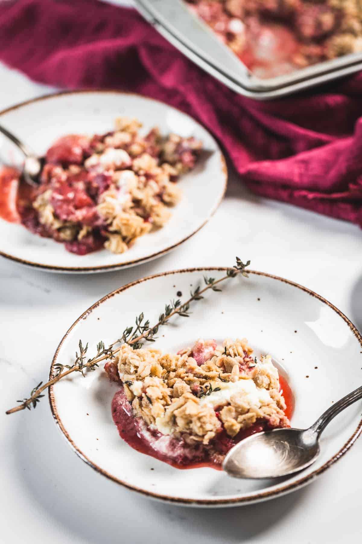 Balsamic Strawberry Crisp With Goat Cheese : At the Immigrant's Table 4 Two white plates each with a serving of Balsamic Strawberry Crisp topped with oat streusel and a sprig of thyme, with silver spoons. A baking dish with more crumble and a red cloth are in the background.