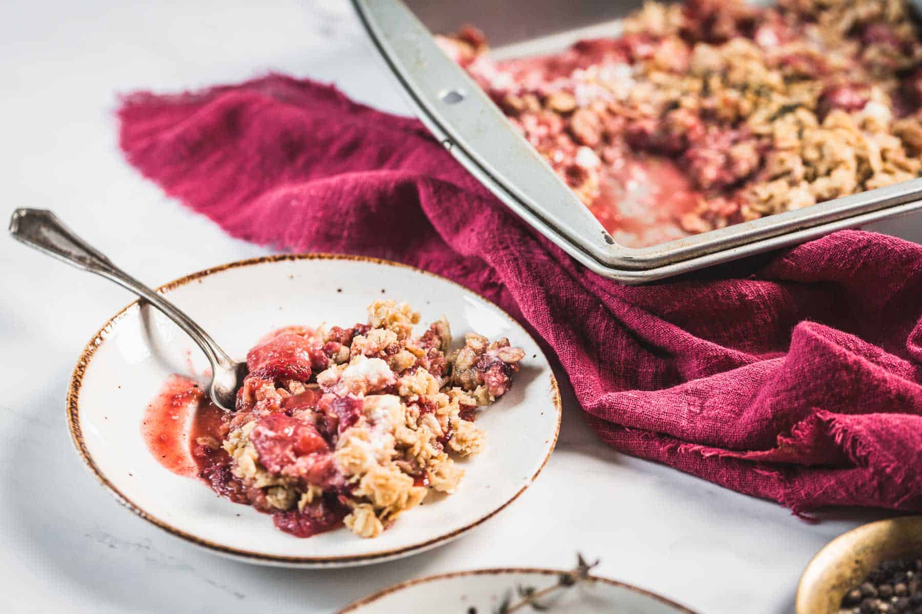 Balsamic Strawberry Crisp With Goat Cheese : At the Immigrant's Table 34 A serving of Balsamic Strawberry Crisp with a silver spoon on a white plate sits next to a baking pan of the same dessert on a white surface with a magenta cloth underneath.