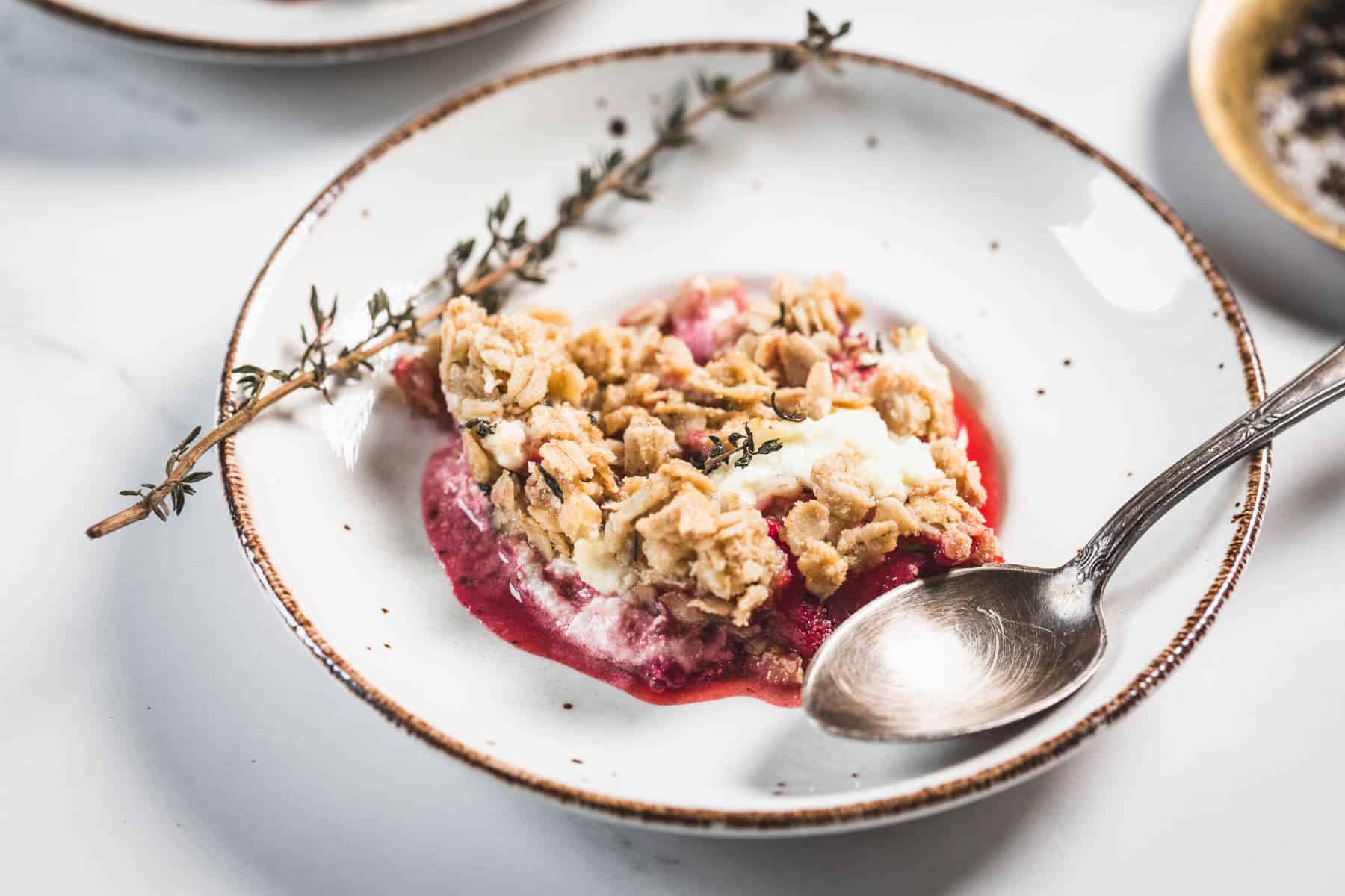 Balsamic Strawberry Crisp With Goat Cheese : At the Immigrant's Table 11 A white plate with a serving of balsamic strawberry crisp topped with oat streusel, some melted ice cream, a sprig of herbs, and a metal spoon resting on the side.