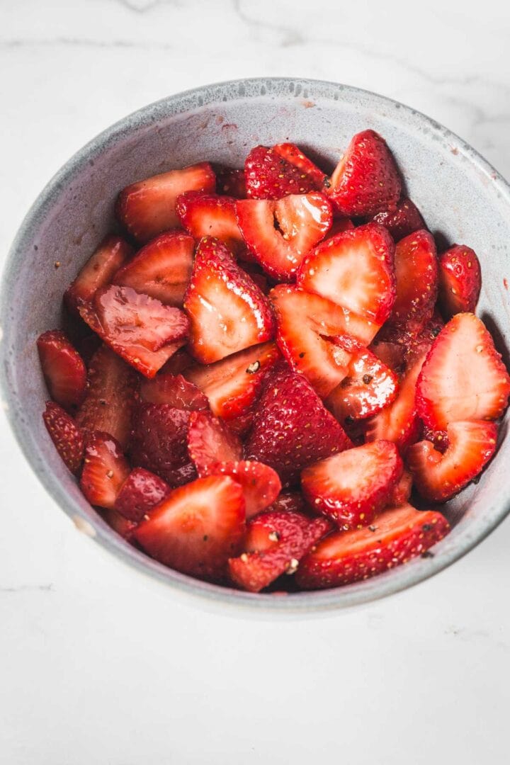 Balsamic Strawberry Crisp With Goat Cheese : At the Immigrant's Table 20 A gray bowl filled with sliced strawberries sits on a white marble surface, perfect for preparing a fresh Balsamic Strawberry Crisp. The strawberries appear vibrant, fresh, and slightly glossy.
