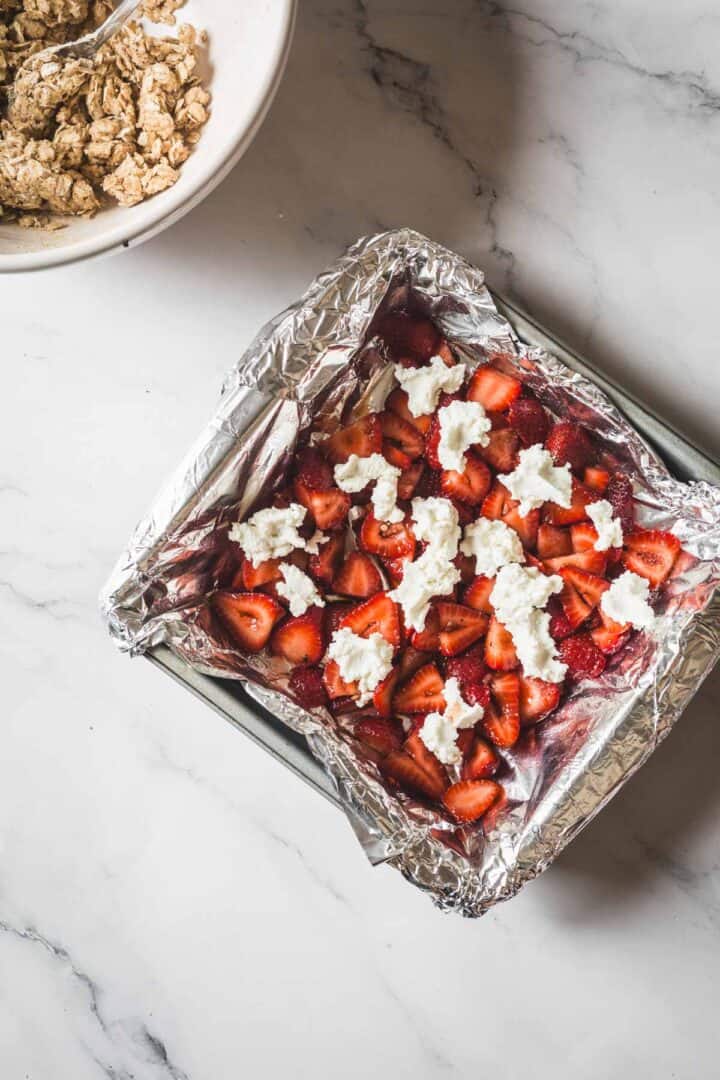 Balsamic Strawberry Crisp With Goat Cheese : At the Immigrant's Table 22 A square baking pan lined with aluminum foil holds sliced strawberries and dollops of white cream or cheese—just the start of a delicious Strawberry Crisp. A bowl with a spoon and crumbly mixture sits nearby on the marble surface.