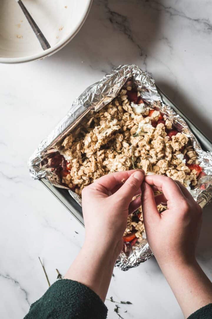 Balsamic Strawberry Crisp With Goat Cheese : At the Immigrant's Table 24 A person is sprinkling a crumbly topping over a foil-lined pan of Balsamic Strawberry Crisp. A mixing bowl with a spoon sits on the white marble countertop, setting the scene for a delicious strawberry dessert.