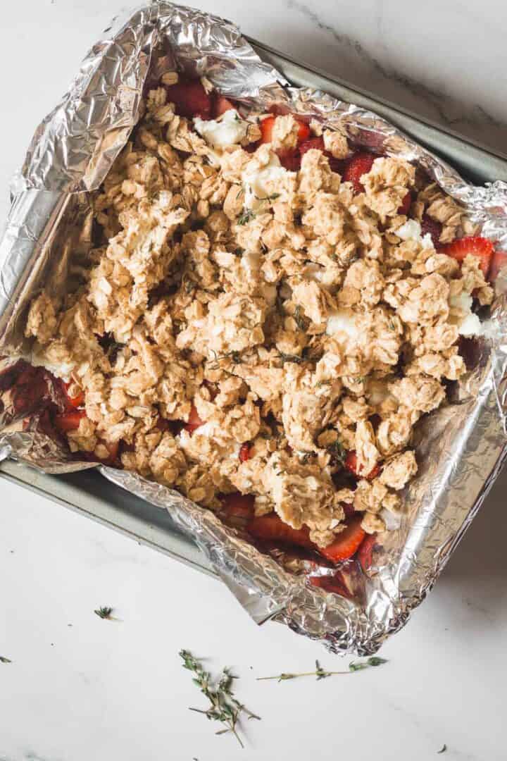 Balsamic Strawberry Crisp With Goat Cheese : At the Immigrant's Table 26 A baking pan lined with foil is filled with sliced strawberries, topped with a crumbly oat mixture for a delicious Strawberry Crisp, sitting on a light-colored countertop. Some fresh herb sprigs are scattered nearby.