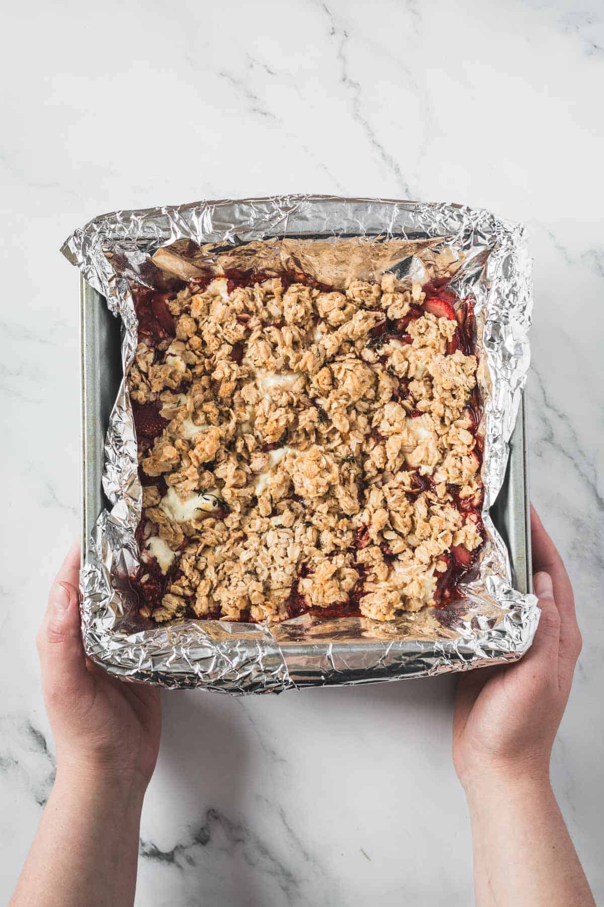 Balsamic Strawberry Crisp With Goat Cheese : At the Immigrant's Table 28 A pair of hands holds a square baking pan lined with aluminum foil and filled with a freshly baked Balsamic Strawberry Crisp, resting on a white marble surface.