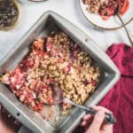 A person scoops Balsamic Strawberry Crisp from a square metal baking pan with a spoon. Some crisp is served on a plate nearby. A red cloth and a small bowl of pepper are also visible on a white surface.