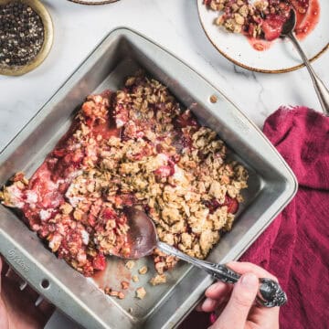 A person scoops Balsamic Strawberry Crisp from a square metal baking pan with a spoon. Some crisp is served on a plate nearby. A red cloth and a small bowl of pepper are also visible on a white surface.