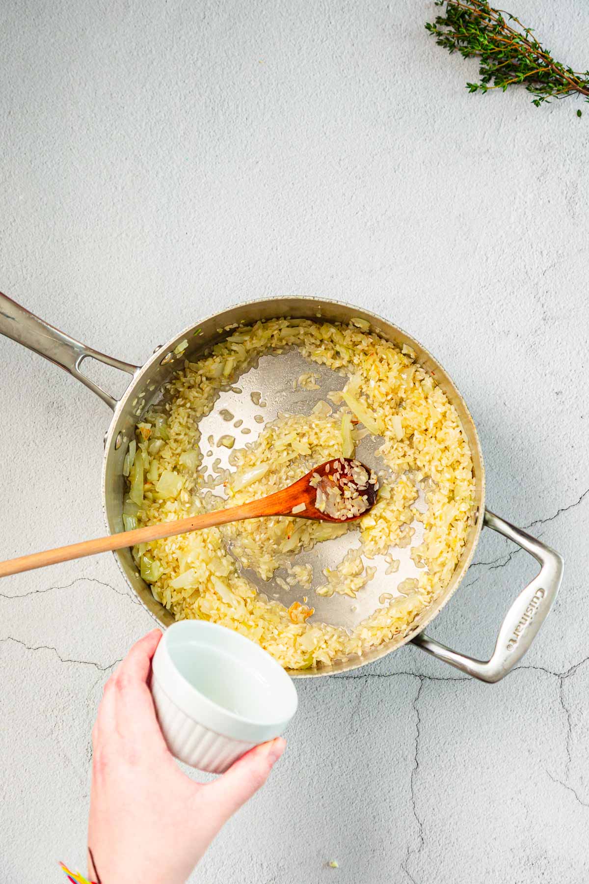 A hand holds a small white ramekin over a metal pan containing sautéed chopped onions. A wooden spoon in the pan holds minced garlic. Fresh herbs lie on the countertop nearby.