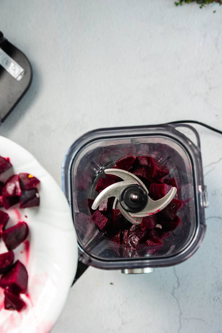 A top-down view of a food processor containing chopped beets. Next to it, a white plate holds additional pieces of chopped beet. The items are placed on a light gray surface.