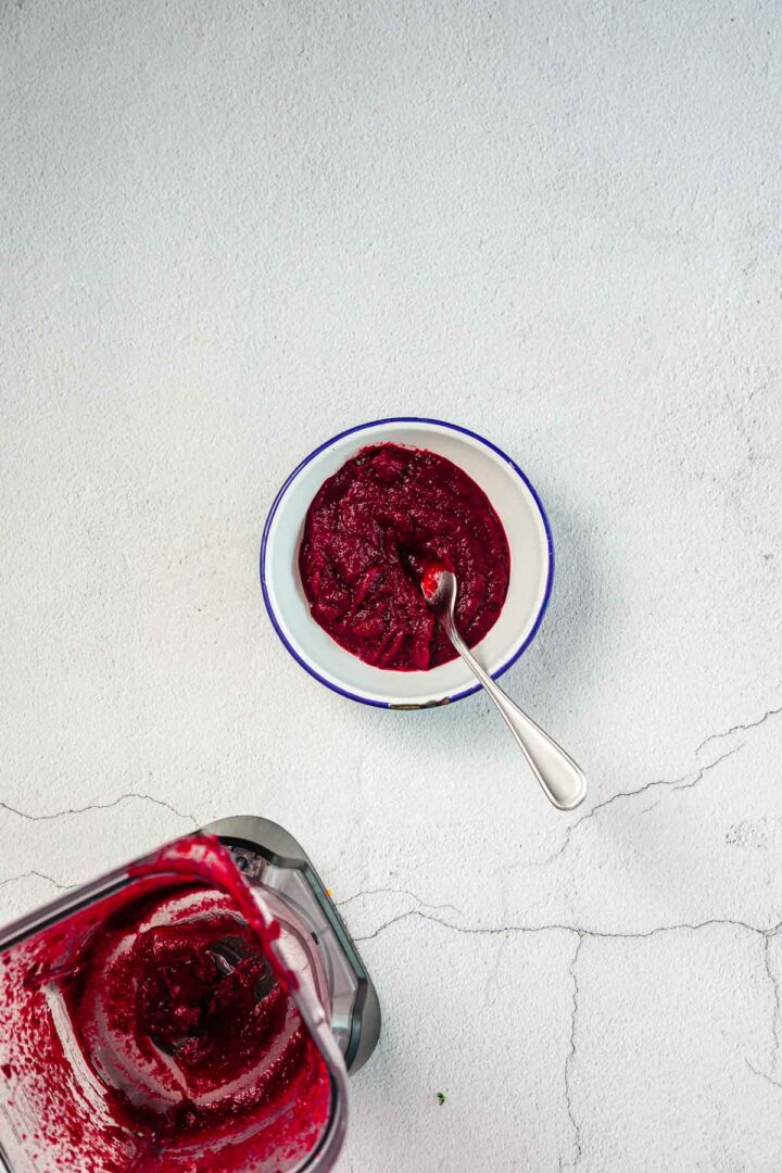 A white bowl with a blue rim contains a thick, dark red sauce or puree with a spoon in it. Below the bowl, a blender with remnants of the same puree is visible on a light gray textured surface.