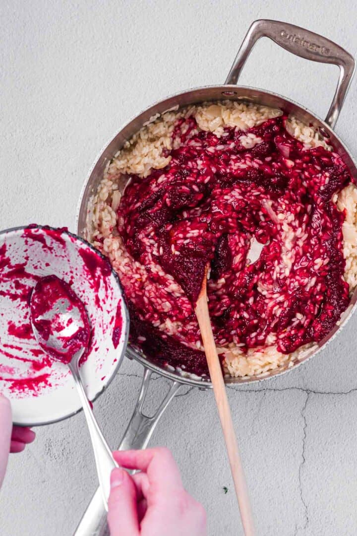 A person stirring a pot of risotto with a wooden spoon, adding a deep red beet mixture from a small bowl. The pan contains partially cooked rice and the mixture is being combined.