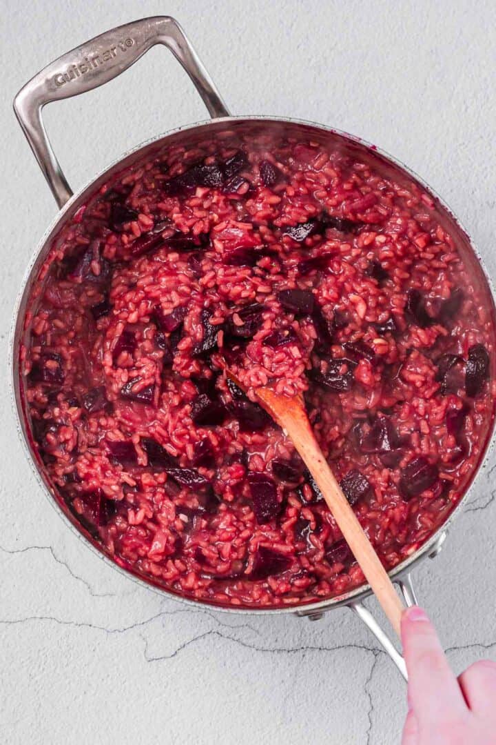 A hand stirs a pot of beetroot risotto with a wooden spoon. The rice and beetroot mixture appears creamy and deep red in color. The pot is on a light-colored surface.