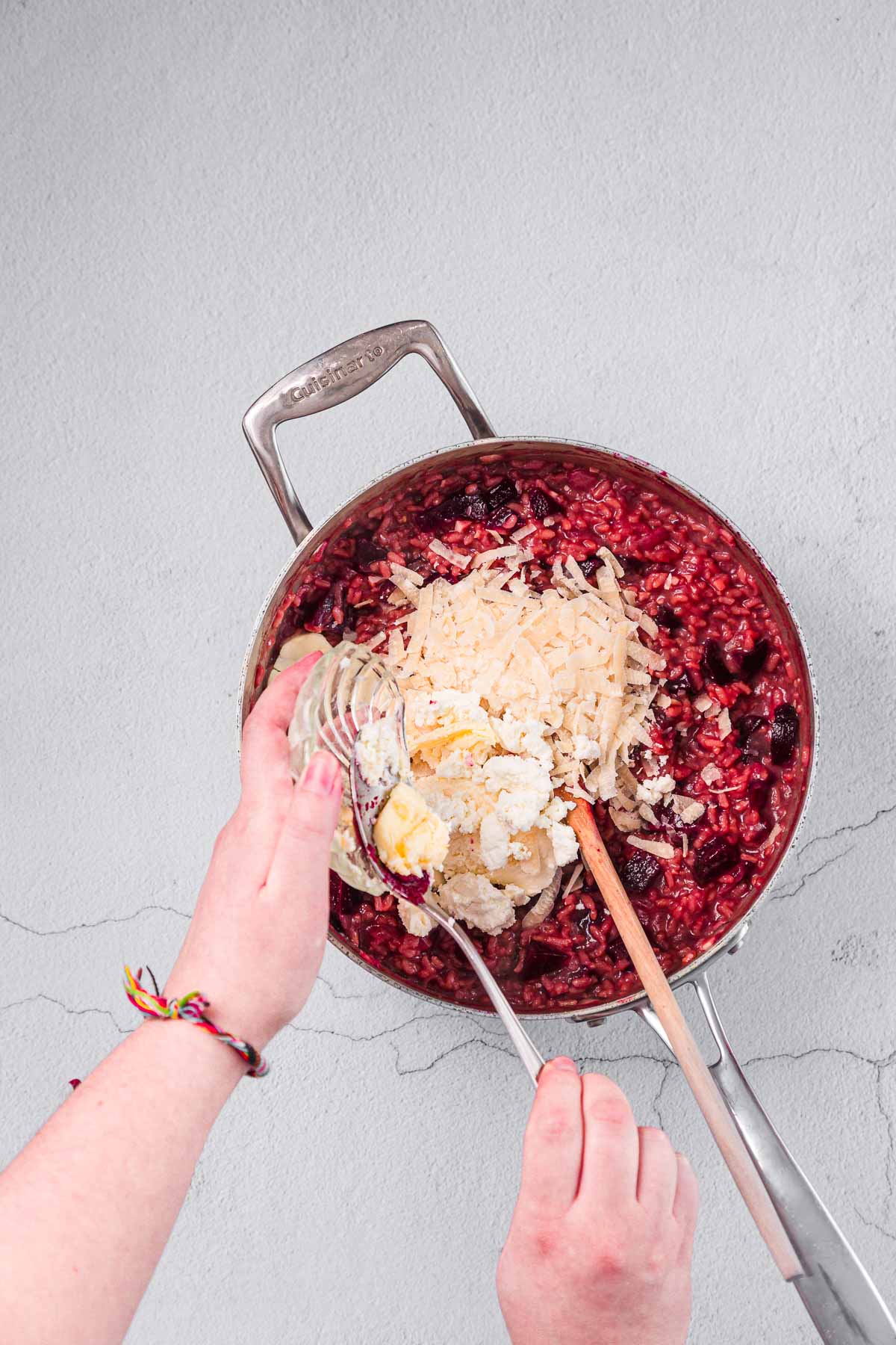 A person adds shredded cheese, butter, and grated cheese to a pan of red risotto with a wooden spoon on a light gray countertop.
