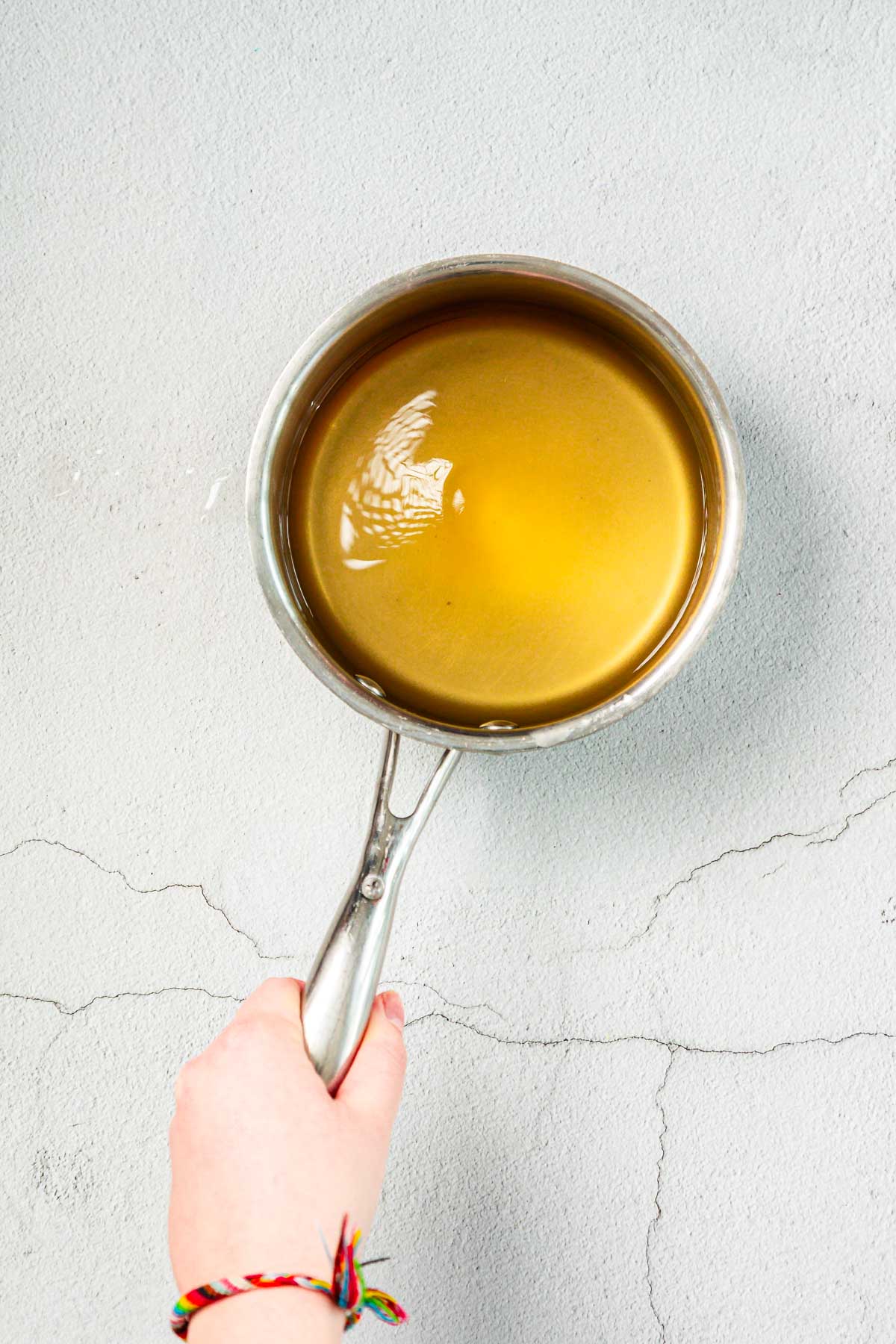 A hand holding a stainless steel saucepan filled with a light brown liquid over a light gray, textured background. The person is wearing a colorful woven bracelet.