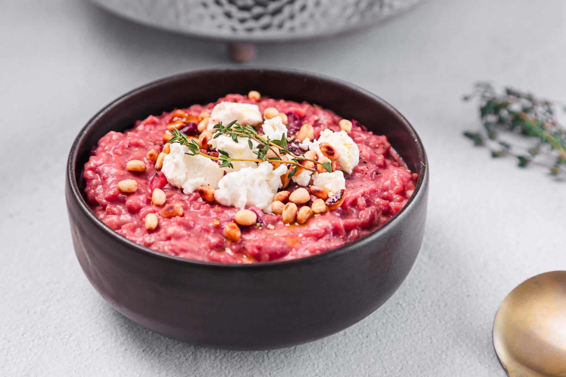 A black bowl filled with beetroot risotto, topped with crumbled cheese, toasted pine nuts, and a sprig of fresh thyme, placed on a light gray surface.