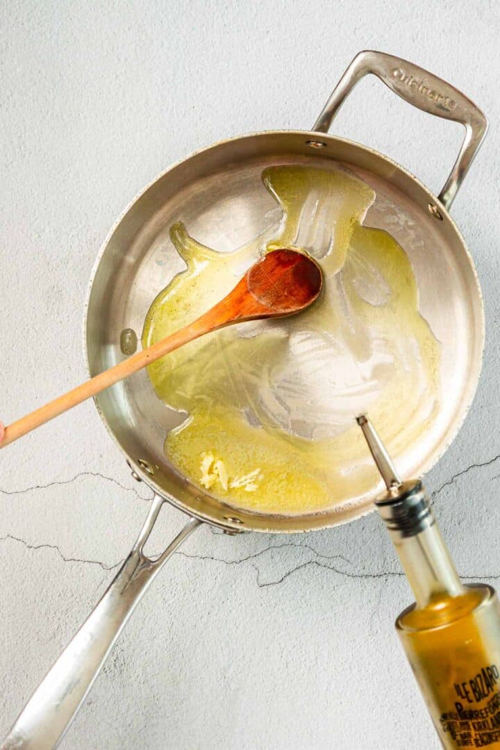 A wooden spoon rests in a stainless steel pan with oil spread inside, while a bottle of olive oil with a pour spout is positioned at the edge of the pan. The pan is on a light-colored surface.